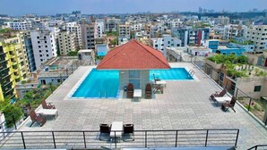 Una piscina en la azotea con techo rojo, rodeada de sillas y mesas de descanso, con vistas a una zona urbana densamente construida.