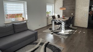 En moderne stue med en grå sofa, et spiseområde med bord og stole, og et køkken i baggrunden.
