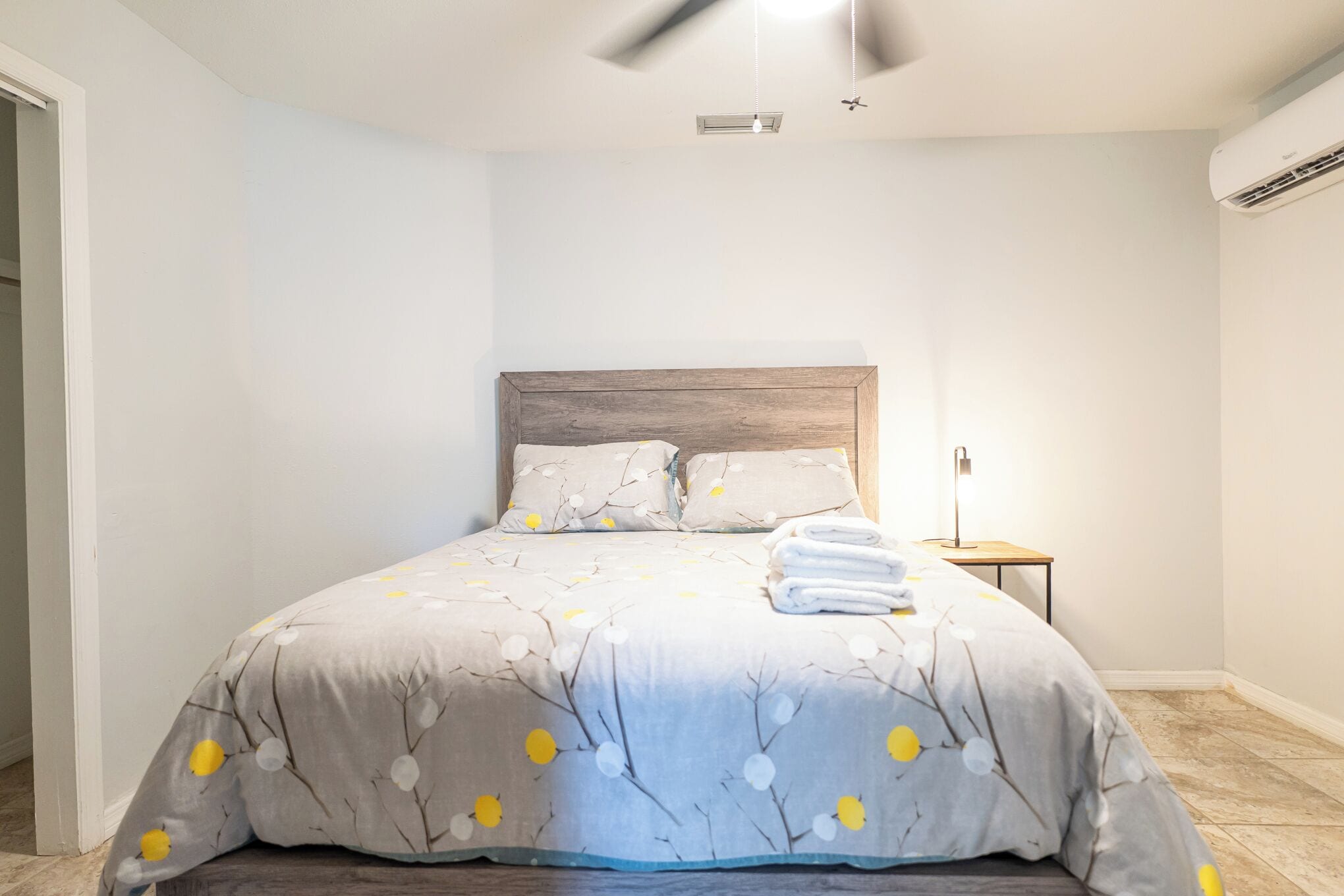 A neatly made bed with a grey floral bedspread, a wooden headboard, and a small bedside table with a lamp.