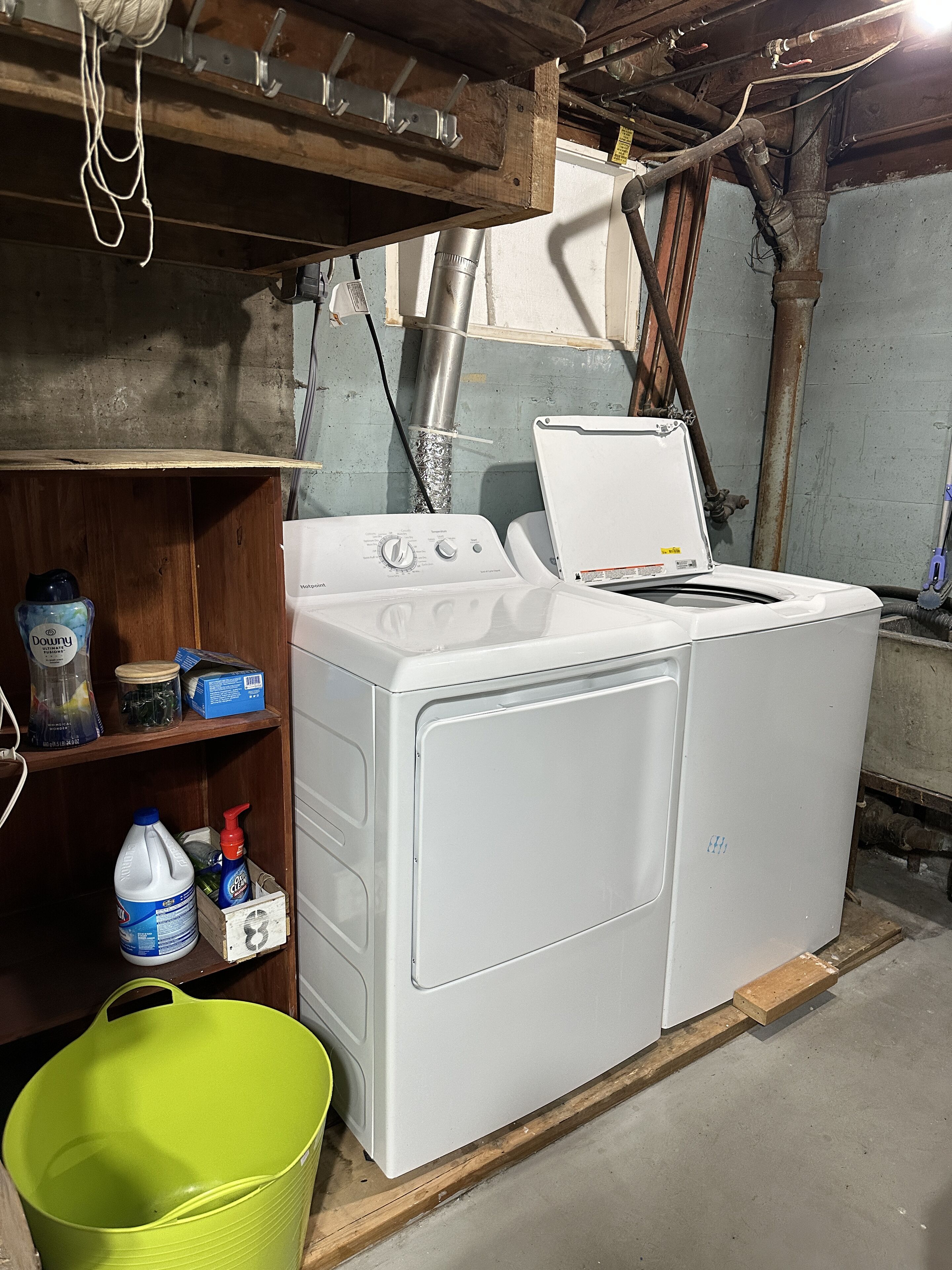 Une buanderie équipée d’un lave-linge et d’un sèche-linge à chargement frontal, d’étagères en bois où sont rangés les produits de nettoyage, et de tuyaux apparents au plafond.