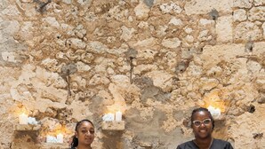 Dos personas de pie en una habitación con paredes de piedra, mesas blancas y velas.