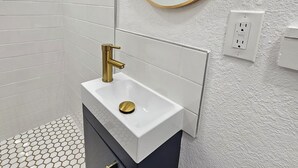 A modern bathroom with a white tiled shower, a gold faucet, a round mirror, and a hexagonal tile floor. - Crashpads (Orlando)
