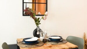 A dining area with a wooden table set for two, two green chairs, a black-framed mirror on the wall, and a vase with flowers on the table.