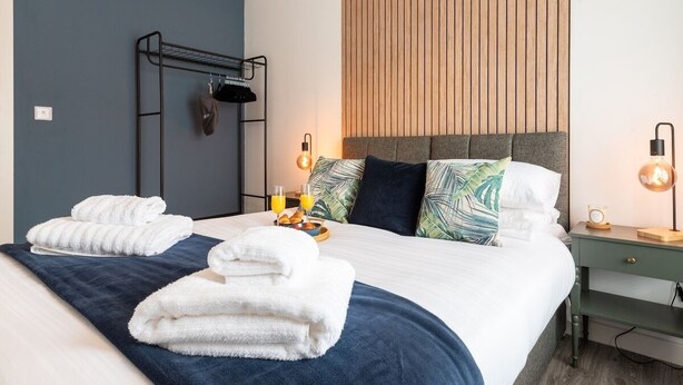 A neatly made bed with towels, a bedside table with a lamp, and a wall-mounted rack with clothes.