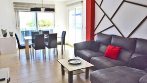 A modern living room with a grey sofa, a dining area with a table and chairs, and a kitchen area.