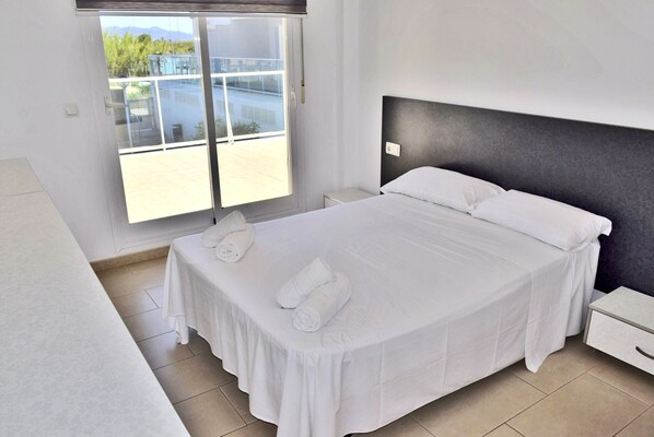 A neatly made bed with white linens and pillows, a bedside table, and a window with a view of greenery and a building outside.
