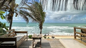 Une terrasse en bois, dotée de bancs et d’une table, offre une vue sur une plage bordée de palmiers et sur l’océan.