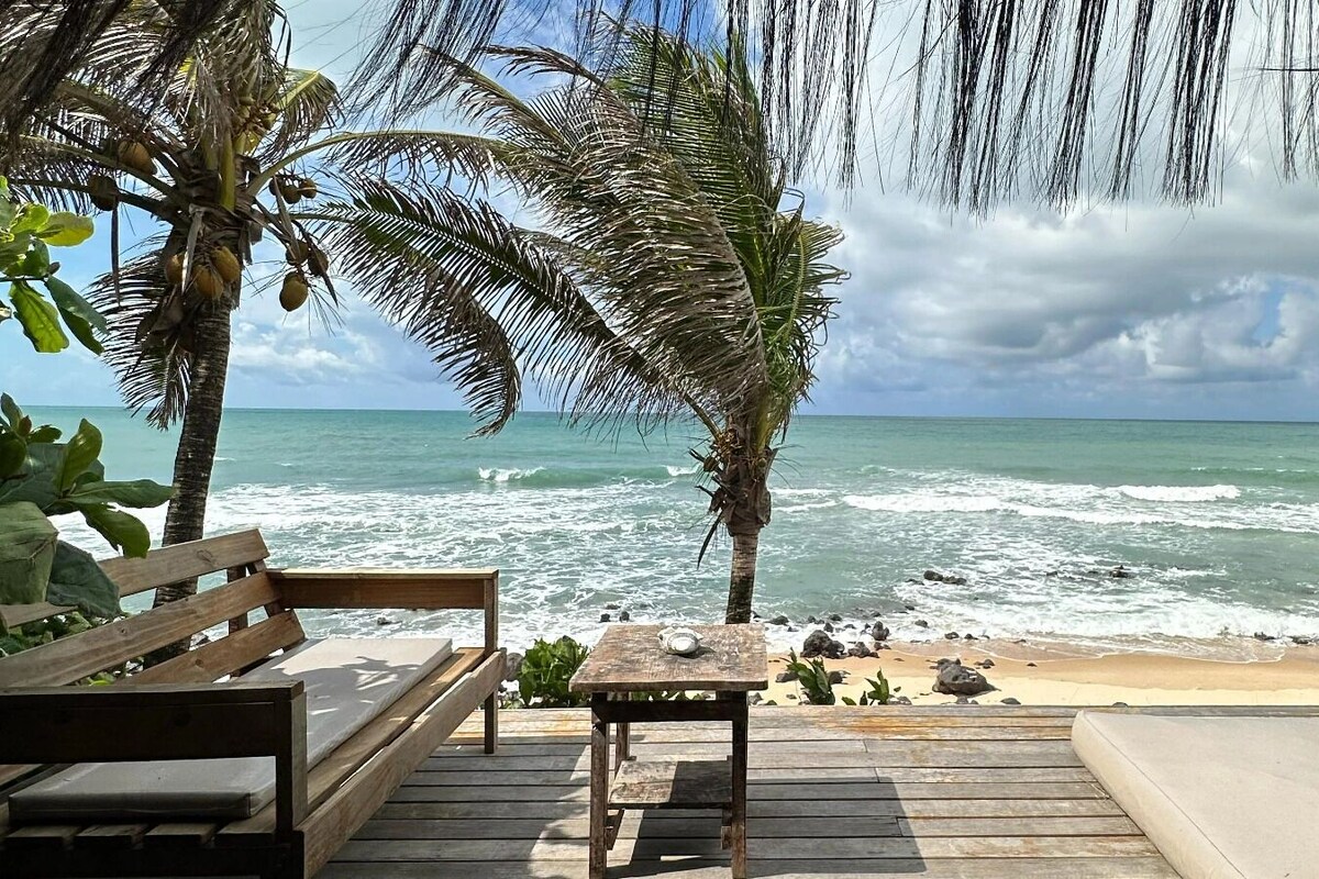 Une terrasse en bois, dotée de bancs et d’une table, offre une vue sur une plage bordée de palmiers et sur l’océan.