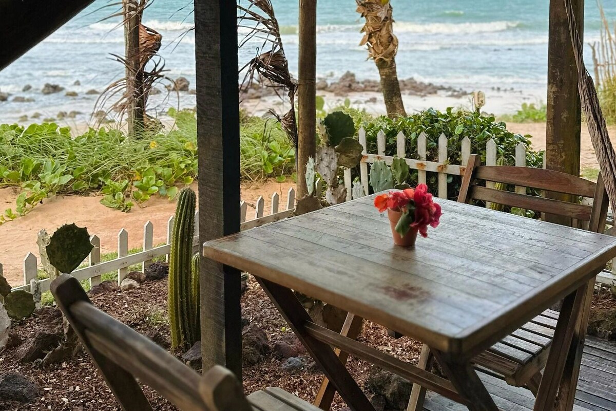 Une table en bois sur laquelle est posée une plante en pot, entourée de cactus et d’une clôture en bois, avec vue sur une plage.