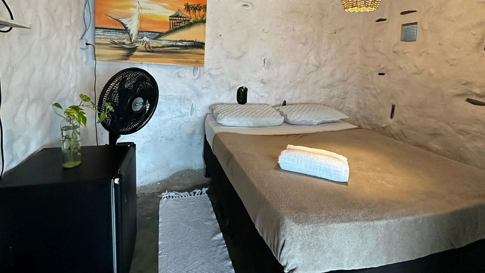 A room with a bed, a fan, a small table with a plant, and a painting on the wall.