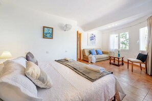 A well-lit room with a bed, a sofa, a small table, and a chair. There are two windows providing natural light.