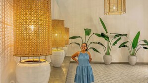 Una mujer con vestido azul parada en un pasillo con piso estampado, lámparas tejidas grandes y plantas en macetas.