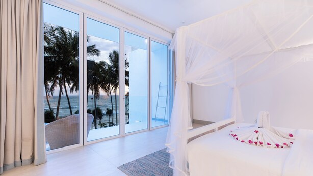 Un dormitorio con un amplio ventanal corredera que da a un balcón con vistas a palmeras y al océano.