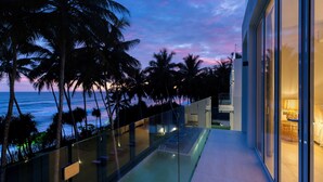 Balcón moderno de hotel con barandilla de vidrio que da a una piscina y palmeras, con vistas al océano y un cielo al atardecer.