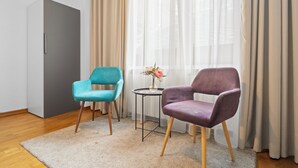 Una habitación con un armario gris, una silla turquesa, una silla morada, una mesita redonda con un jarrón de flores y cortinas de gasa.