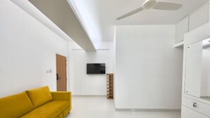 A modern living room with a yellow sofa, a ceiling fan, a flat-screen TV mounted on the wall, and a wooden cabinet. - 600sqft,5 Elite Stay with Pool, Sauna and Gym in Bashundhara (Dhaka)