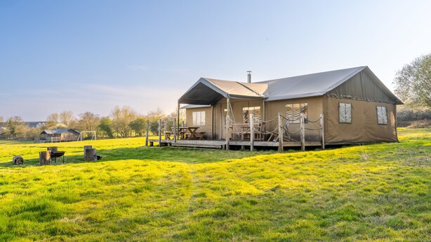 A rustic cabin with a porch and outdoor seating area, surrounded by a grassy field and trees. - Dog Friendly Safari Tent Cuckoo at Sweffling Hall, Suffolk (Sweffling)