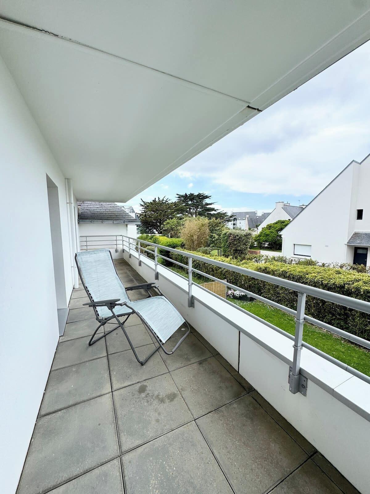 Ein Balkon mit einem Metalltisch und -stühlen, einem weißen Geländer und Blick auf eine Wohngegend mit Häusern und Grünflächen.