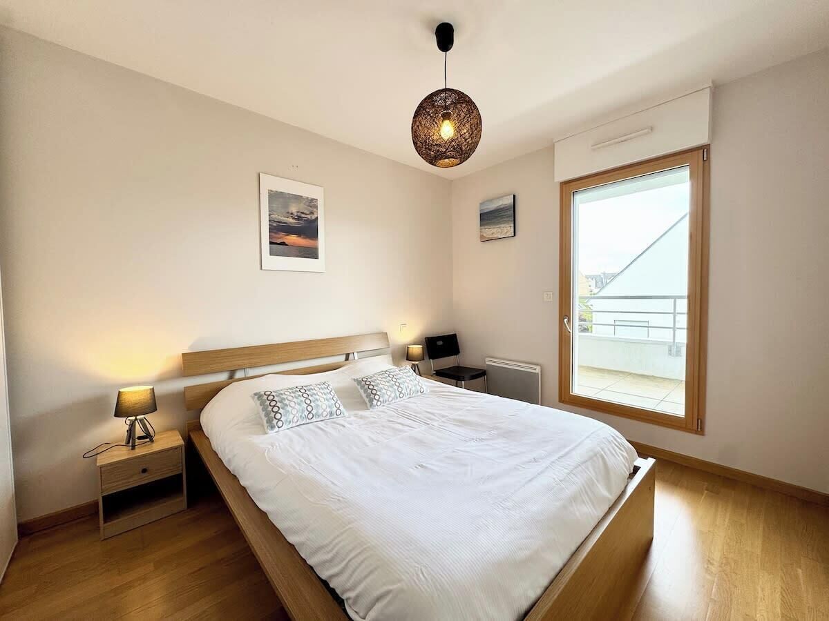 A bedroom with a large bed, a bedside table with a lamp, a radiator, and a window with a view of a balcony.