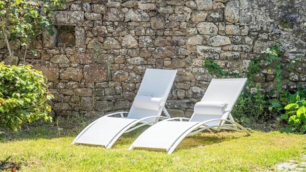 Two white lounge chairs with metal frames, positioned on a grassy area in front of a stone wall.