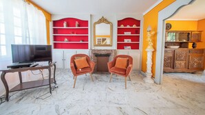 A living room with a TV, two armchairs, a fireplace, red shelves, and a kitchen area.