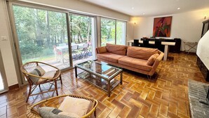 Amplia sala de estar con un gran ventanal corredero que da a un patio exterior, un sofá marrón, una mesa de centro de vidrio y sillas de mimbre.