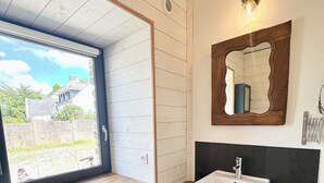 Une salle de bain avec une fenêtre, un lavabo, un miroir et une petite poubelle.
