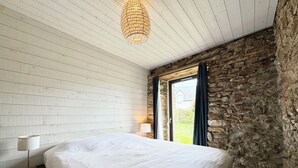 Une chambre à coucher avec un lit, une table de chevet, une lampe, un mur en pierre et une fenêtre avec des rideaux.
