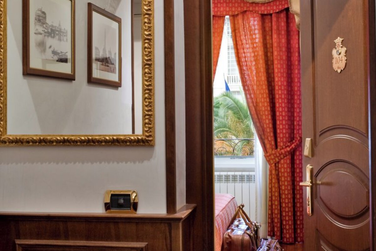A hotel room with a wooden door, a mirror with framed pictures, and a view of the outside through a window with red curtains.