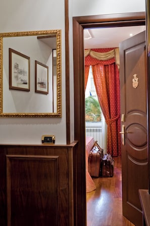A hotel room with a wooden door, a mirror with framed pictures, and a view of the outside through a window with red curtains. - De Luxe Inn 6 Guest House (Rome)
