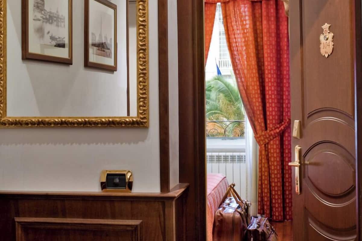 Une chambre d’hôtel avec une porte en bois, un miroir orné de cadres et des photos, et une vue sur l’extérieur par une fenêtre avec des rideaux rouges.