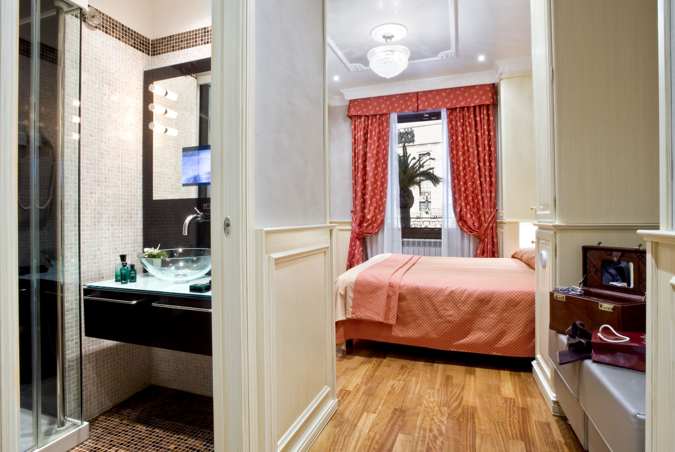 A hotel room with a glass shower enclosure, a bed with a red bedspread, a vanity with a mirror, and a small seating area with a chair and a table.