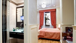 A hotel room with a glass shower enclosure, a bed with a red bedspread, a vanity with a mirror, and a small seating area with a chair and a table. - De Luxe Inn 6 Guest House (Rome)