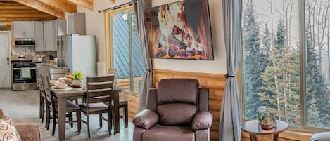 A cozy living room with a fireplace, wooden ceiling beams, a dining area, and a view of trees through large windows.