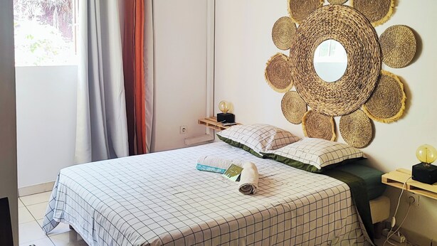 Une chambre avec un plafond en bois, un lit recouvert d’une courtepointe à carreaux, une table de chevet avec une lampe et un miroir tissé fixé au mur.