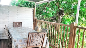 Une terrasse extérieure couverte, avec une table et des chaises en bois, entourée d’une clôture en bois et d’une végétation luxuriante.