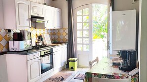 Une cuisine avec des meubles blancs, un four à micro-ondes, une cuisinière et un réfrigérateur. Il y a une fenêtre avec des rideaux, un petit tabouret et une table avec une chaise.