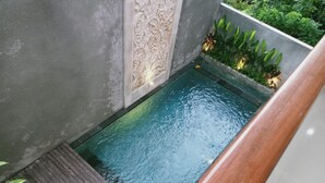 Une petite piscine rectangulaire entourée d’une terrasse en bois, adossée à un mur gris orné de panneaux décoratifs.
