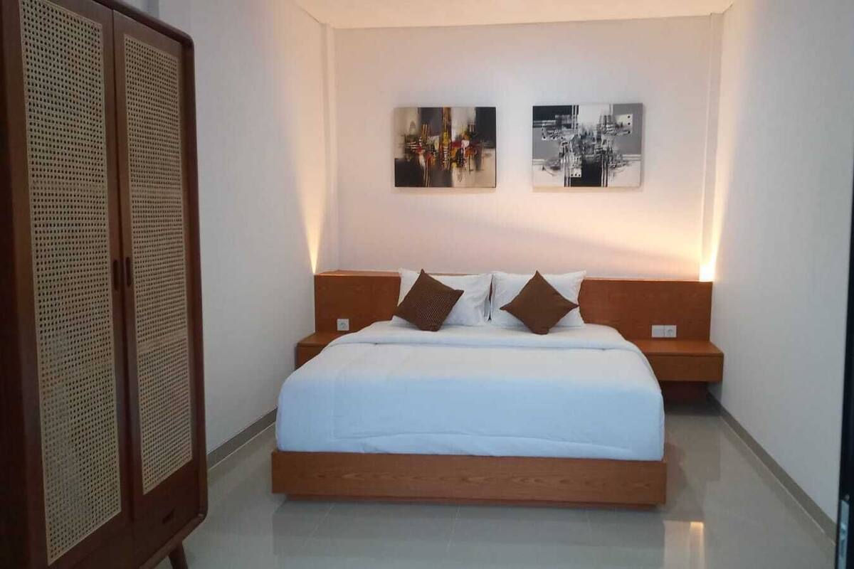 Une chambre à coucher avec un lit en bois, une literie blanche, deux œuvres d’art accrochées au mur et une armoire en bois.