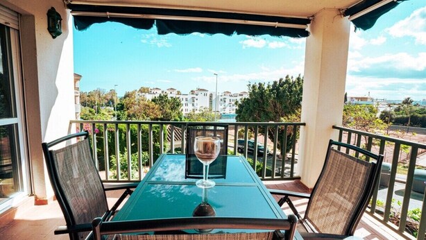 Een balkon met tafel en stoelen, een glas wijn en uitzicht op gebouwen en groen.