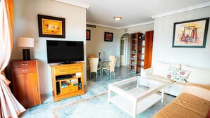 Een woonkamer met een flatscreen televisie, een witte bank, een salontafel, een houten kast en een eethoek met een tafel en stoelen.