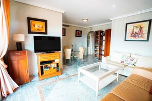 A living room with a flat-screen TV, a white sofa, a coffee table, a wooden cabinet, and a dining area with a table and chairs. - Locrimar, 3 bedroom apartment in Puerto Banus (Marbella)
