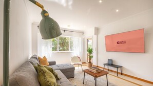 A modern living room with a grey sofa, a coffee table, and a green floor lamp. There is a large pink sign on the wall and a window with a view of trees.