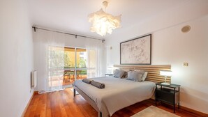 A bedroom with a bed, bedside tables, a hanging light fixture, a window with curtains, and a painting on the wall.