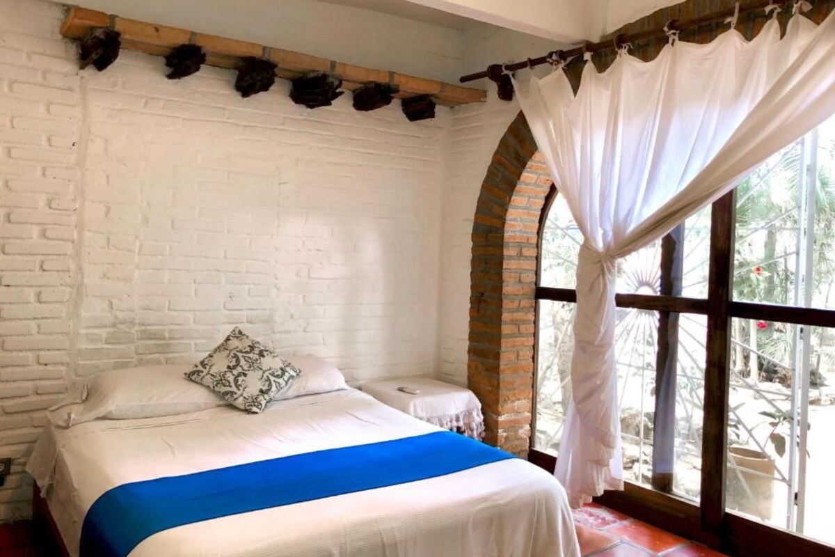 A room with a bed, white brick walls, a window with curtains, and a view of greenery outside.