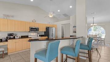 A modern kitchen with wooden cabinets, a stainless steel refrigerator, and a bar area with blue chairs.