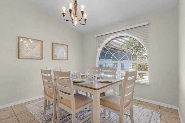 A dining area with a wooden table, six chairs, two framed artworks, and a chandelier. - Sunny Cape Coral 4BR Heated Pool & Screened Lanai (Cape Coral)