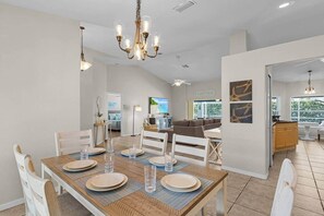 A dining area with a wooden table set for four, a chandelier, and a view of the living room and kitchen in the background. - Sunny Cape Coral 4BR Heated Pool & Screened Lanai (Cape Coral)