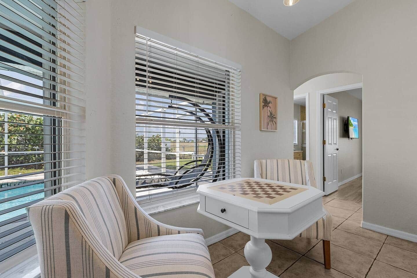 A room with a striped chair, a small table with a chessboard, and a window with blinds.
