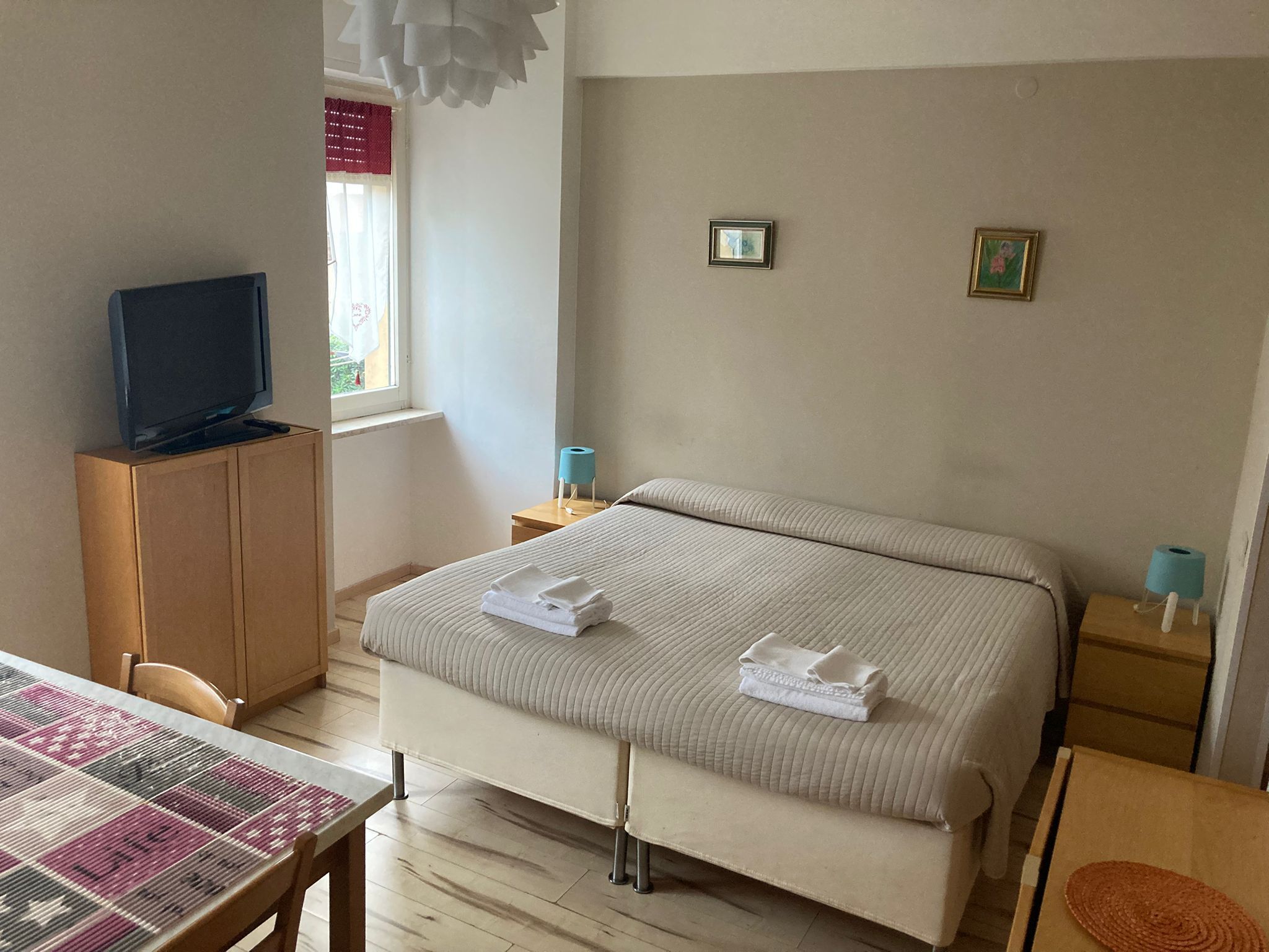 A hotel room with a bed, a television on a cabinet, a table with a patterned tablecloth, and a window with curtains.